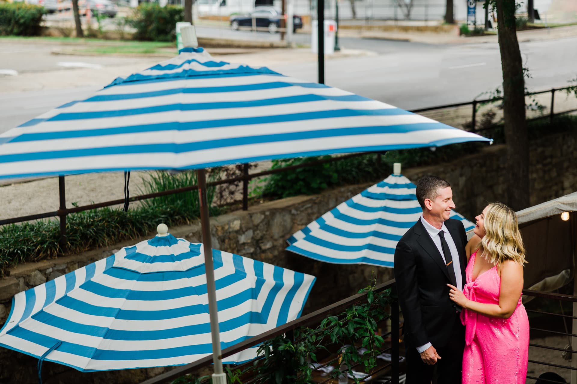 A couple embraces with the bride in a bold pink sequin dress, the contrasting backdrop of blue and white striped umbrellas makes them pop within the photo, the couple is smiling and laughing at eachother.