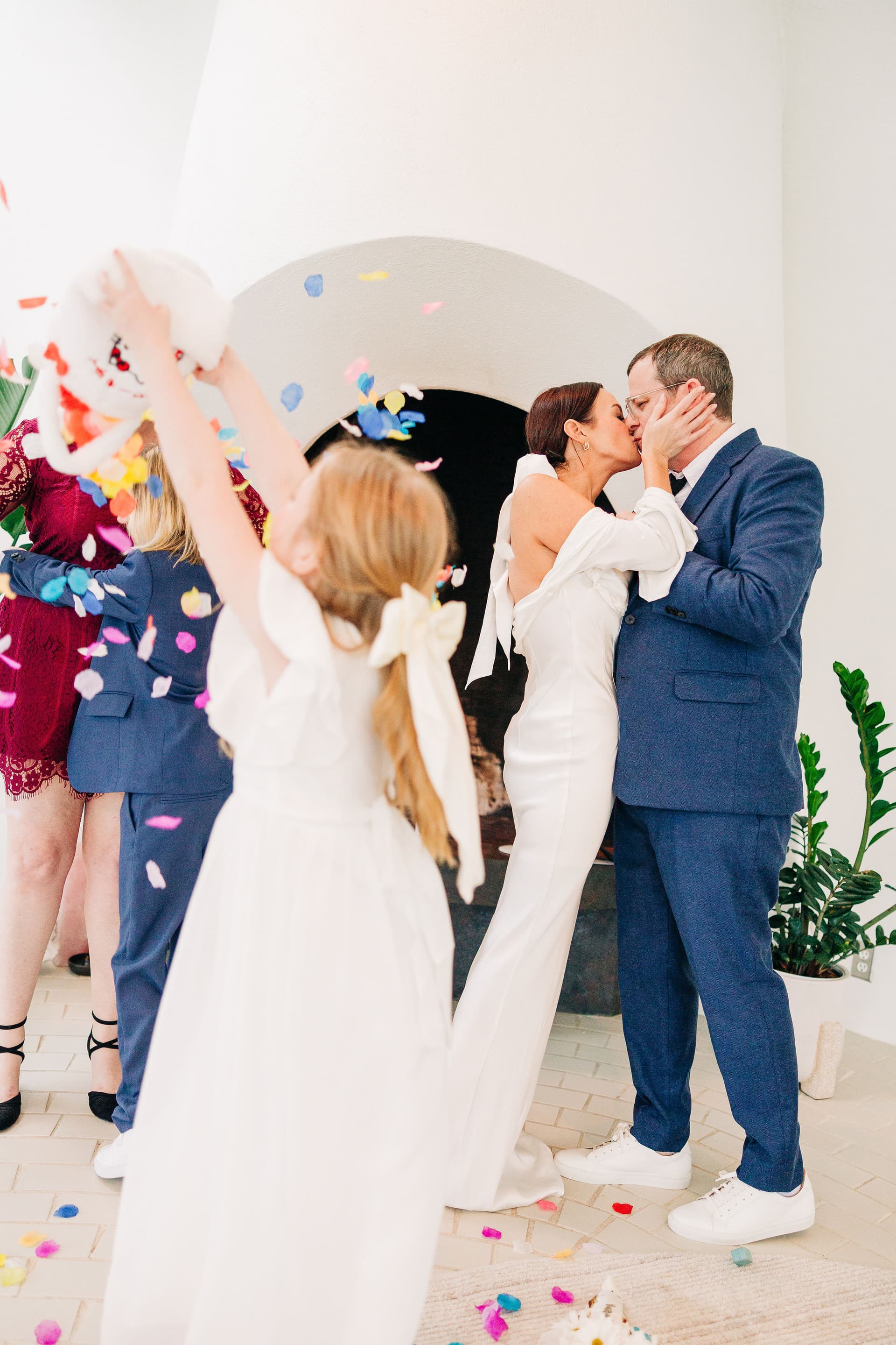 A couple shares their first kiss as the flower girl dumps the flowers on the ground in the foreground of the frame. They are in an intimate airb&b on the lake with natural light.