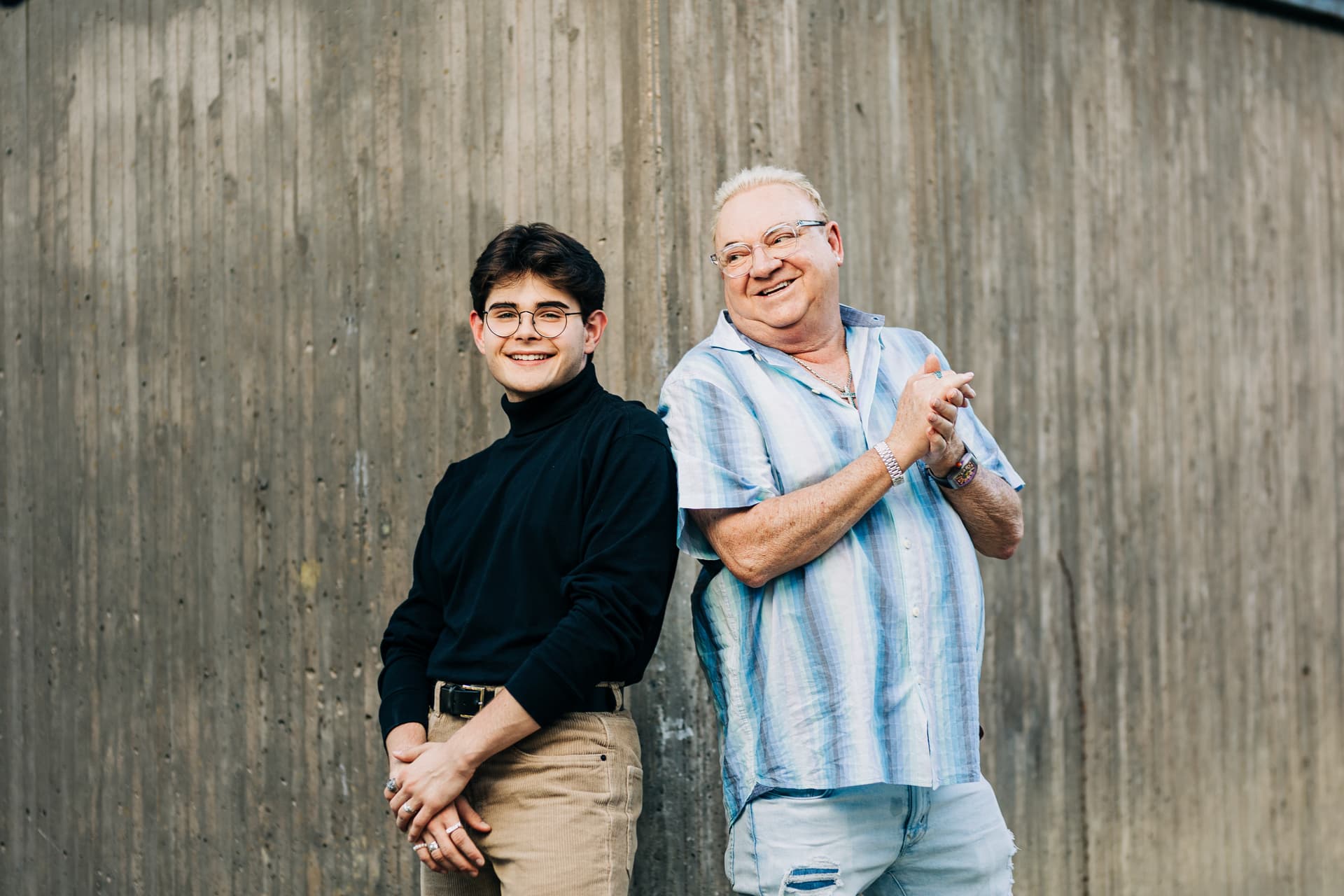 A father and son stand back to back against a modern concrete wall, both smiling candidly with the father looking towards the son.