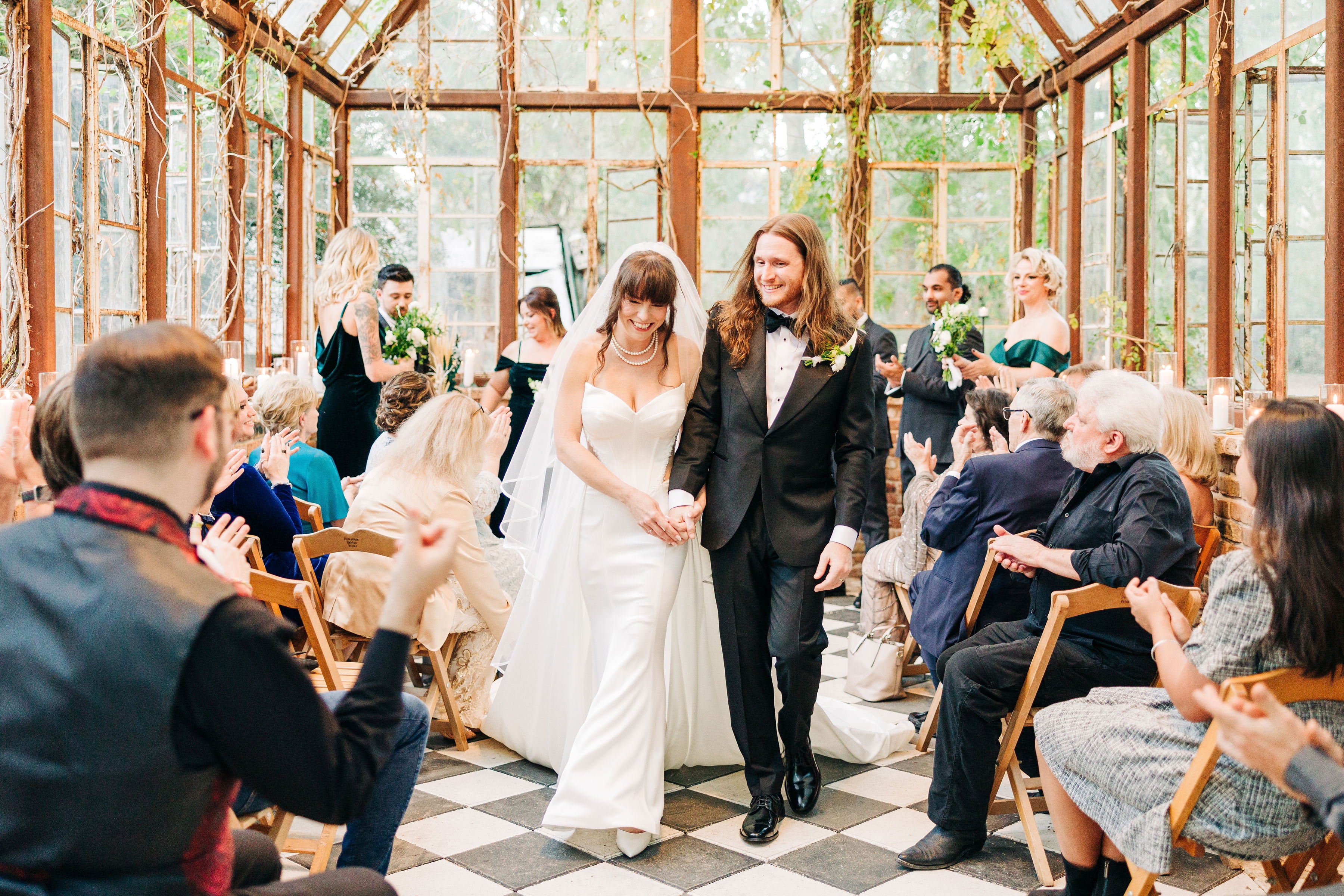 A couple walks down the aisle after the ceremony, smiling as friends within the greenhouse of Sekrit Theater clap for them.