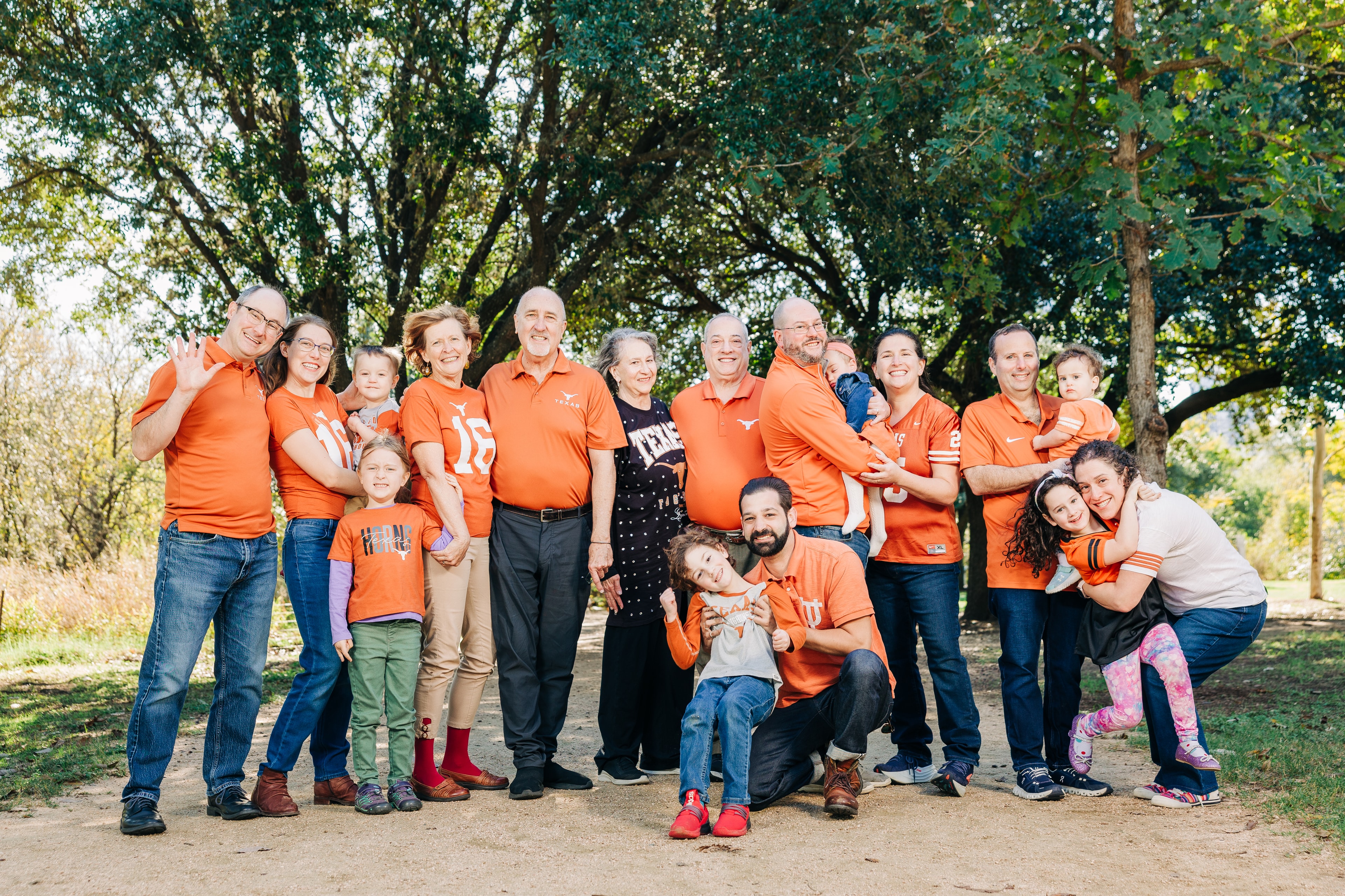 Three generations of a family posing together in coordinated UT Austin themed outfits, smiling towards the camera with oak trees in the background and candid posing.