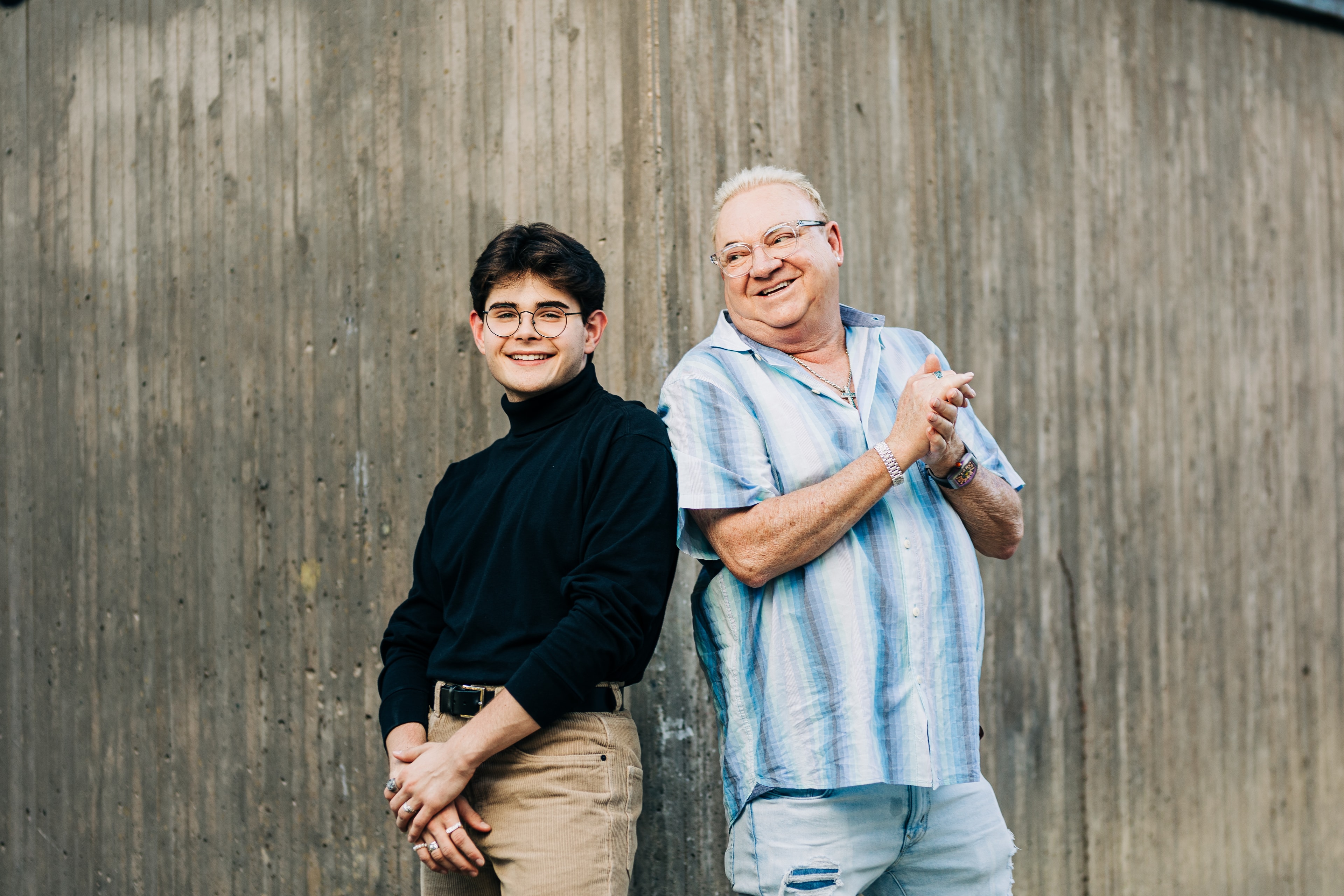 A father and son stand back to back against a modern concrete wall, both smiling candidly with the father looking towards the son.