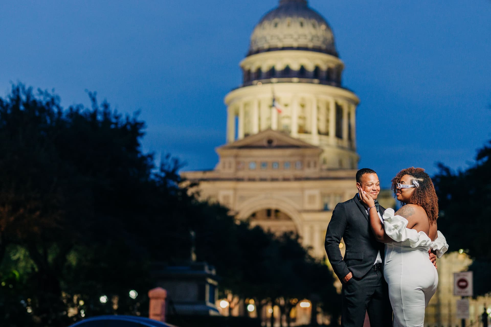 A couple in front of the Texas State Capitol, dressed in wedding attire lit with studio lighting, with blue-hour lighting behind them. The capitol is lit up with warm orange light.