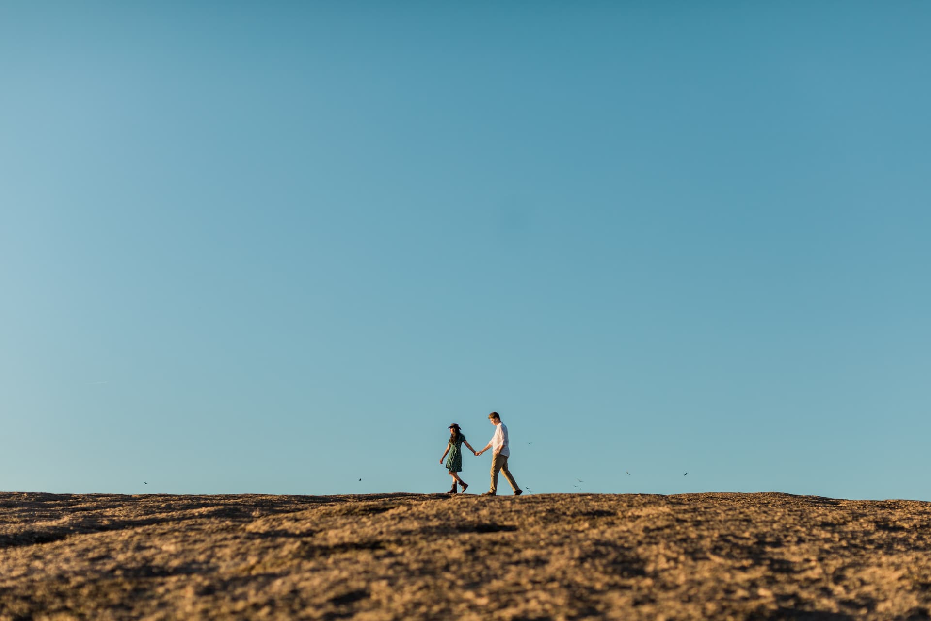 Couple walking across the stone surface of Enchanted Rock. The wide landscape and blue skies surround them as birds fly in the distance.