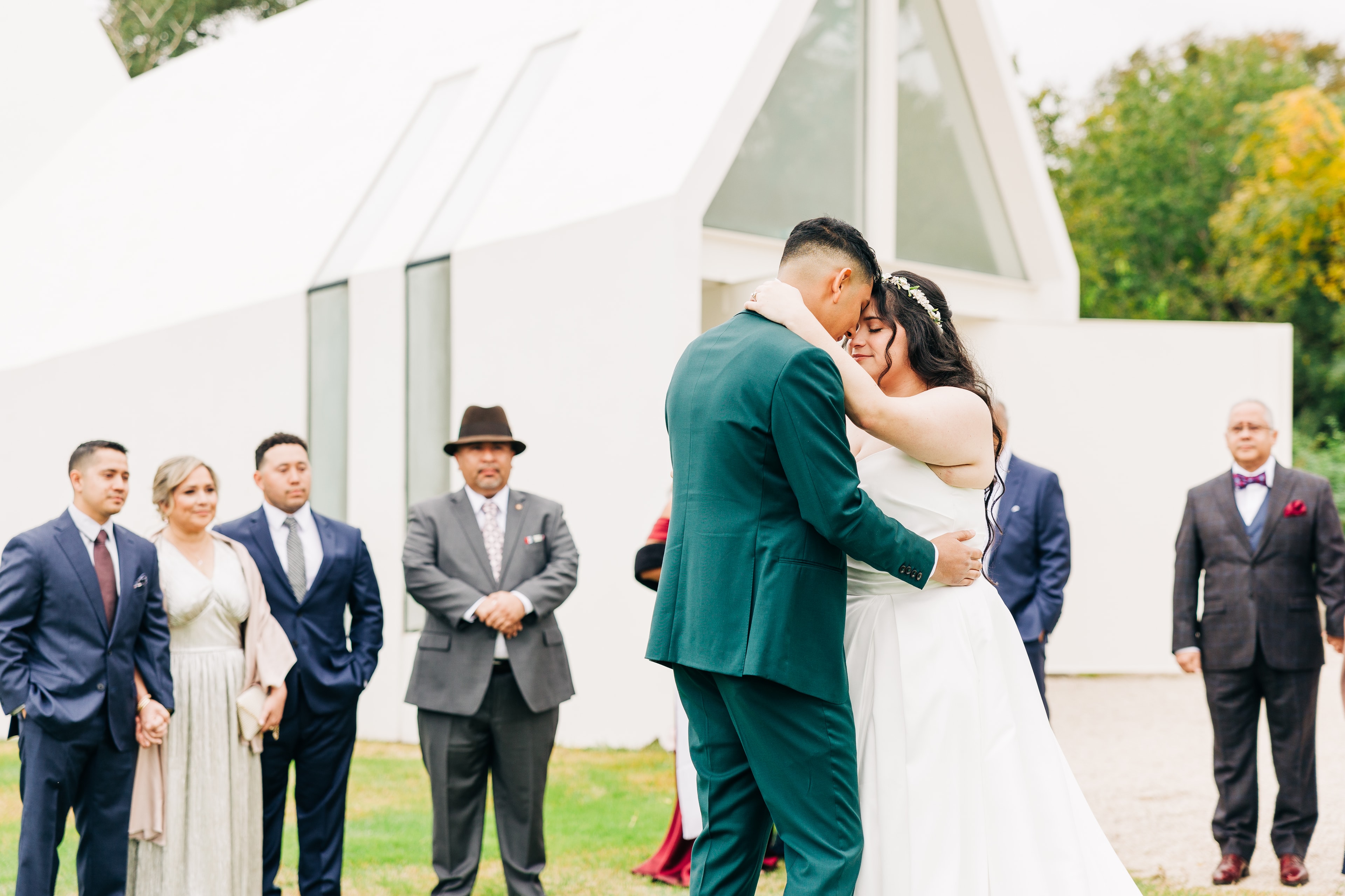 A bride and groom share their outdoor first dance as their family is in a circle around them. The scene is full of emotion.