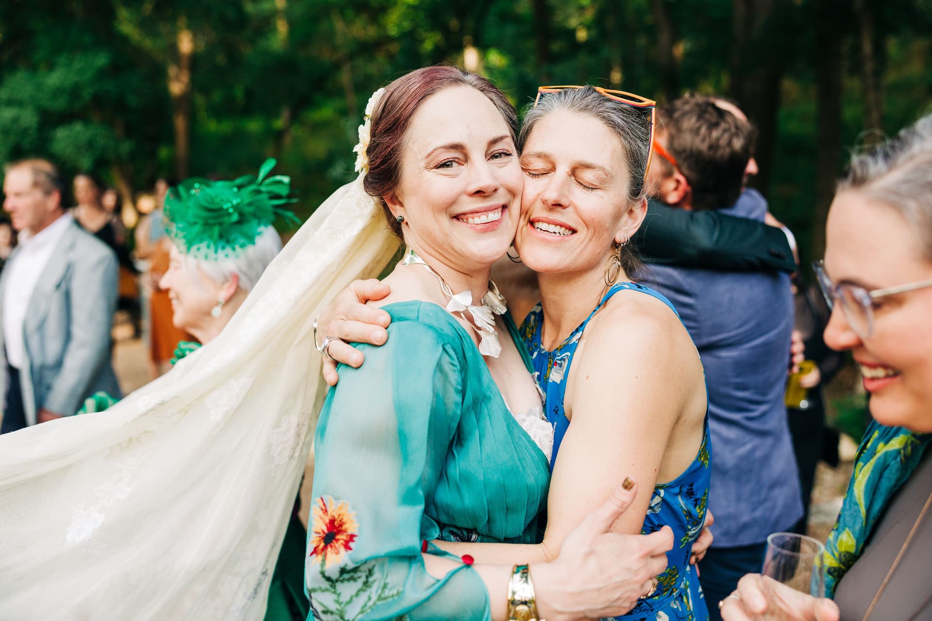 A bride shares a warm embrace after the ceremony with a guest, her veil flowing in the wind with natural light and a crowd around them.