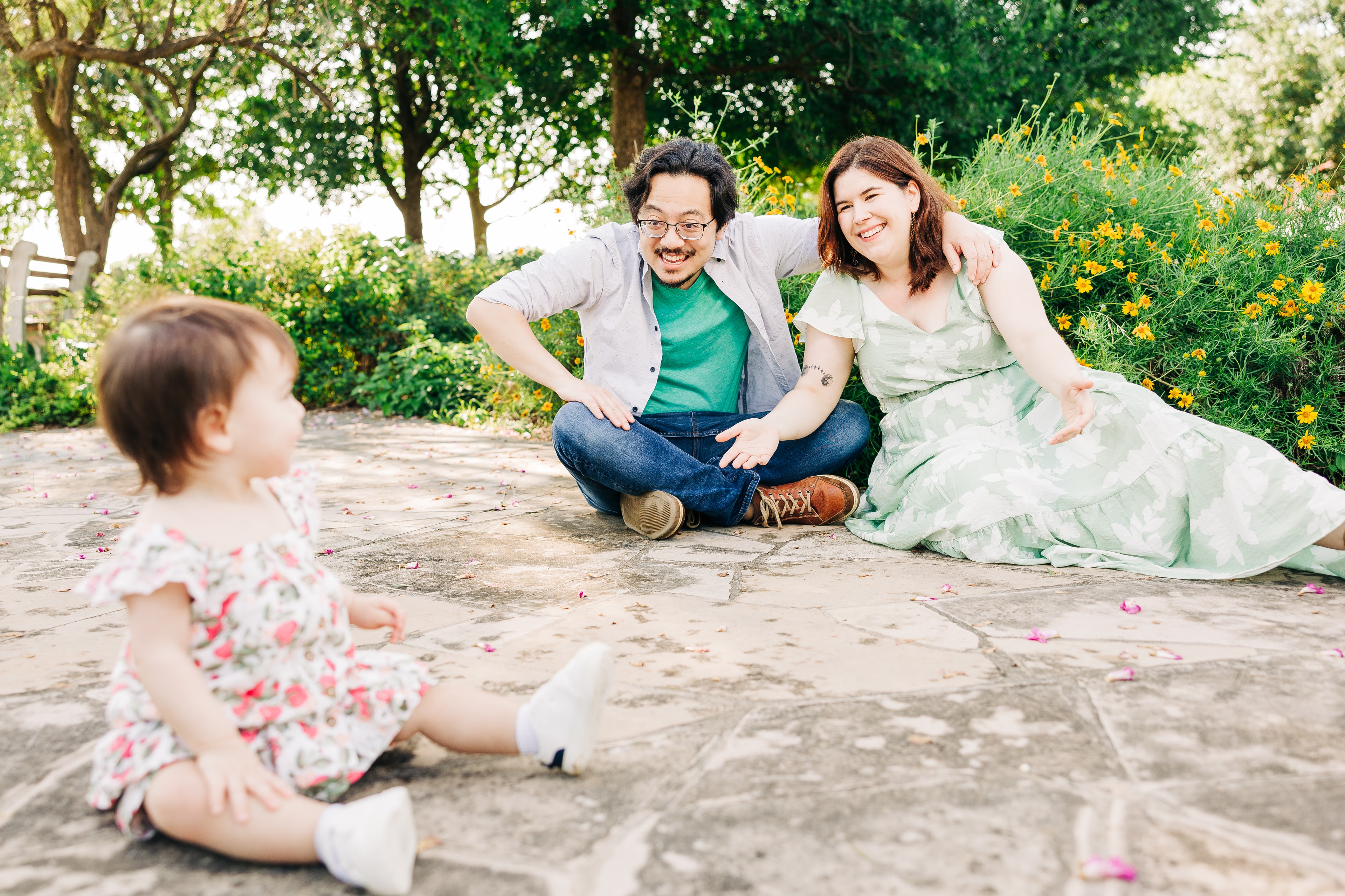 A couple smiles joyfully towards their toddler sitting on the pathway of the Butterfly Gardens, with golden hour lighting.