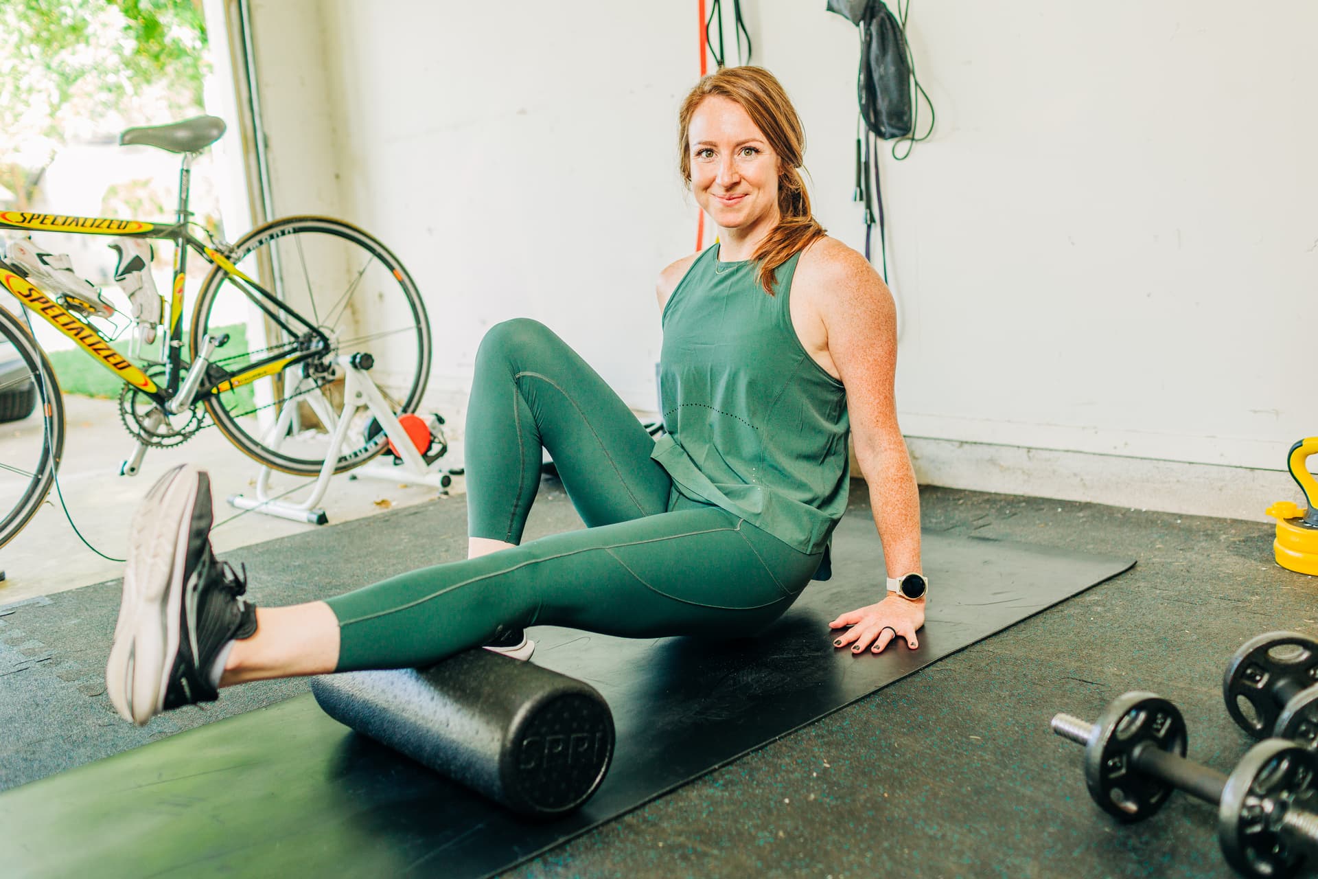 A self employed fitness coach in a natural, candid environment at her home gym. she is foam rollin her leg with a soft, inviting smile.