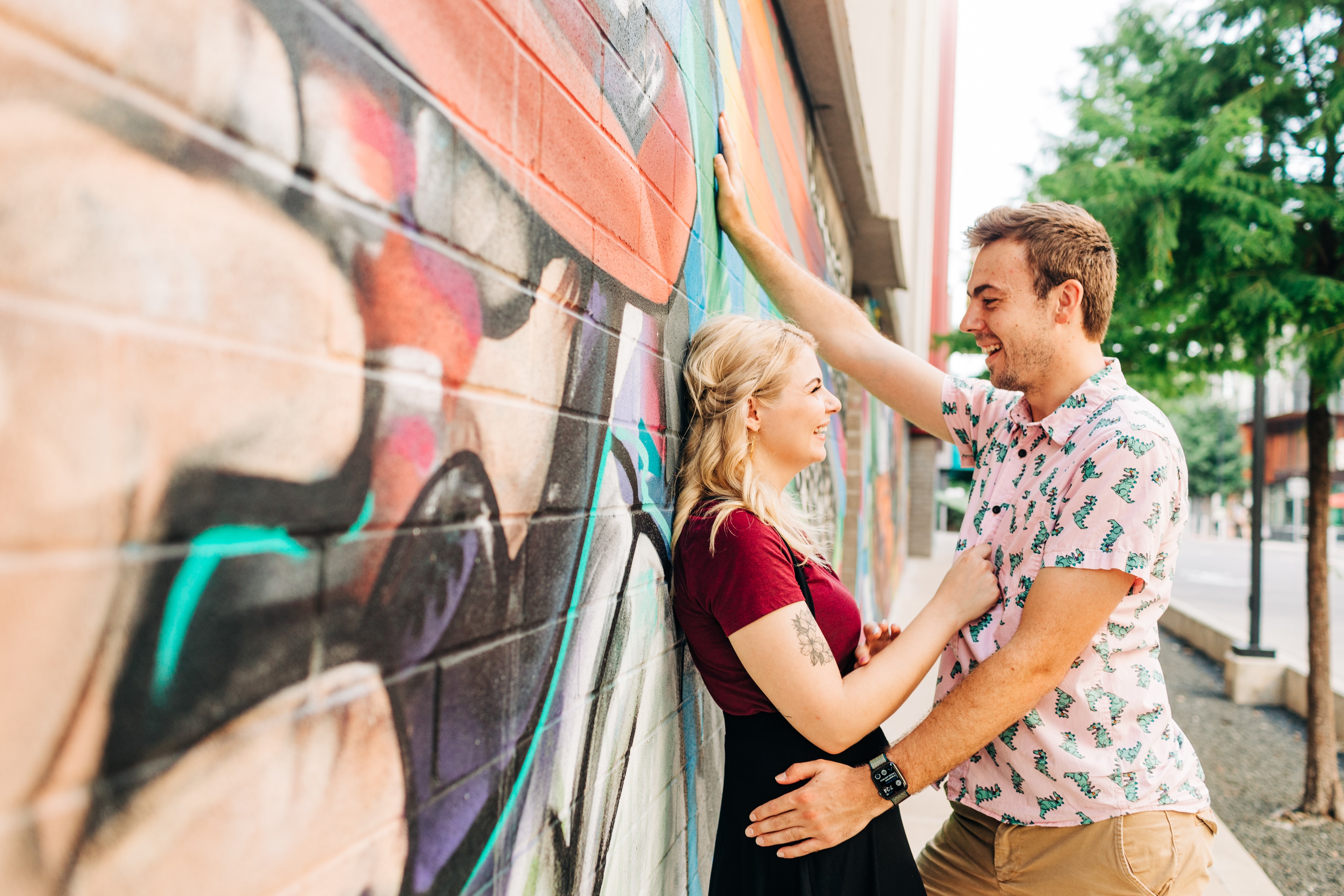 Couple cuddled up to eachother against a graffiti wall near the South Congress Alamo Drafthouse area, the lighting is natural and their smiles are authentic