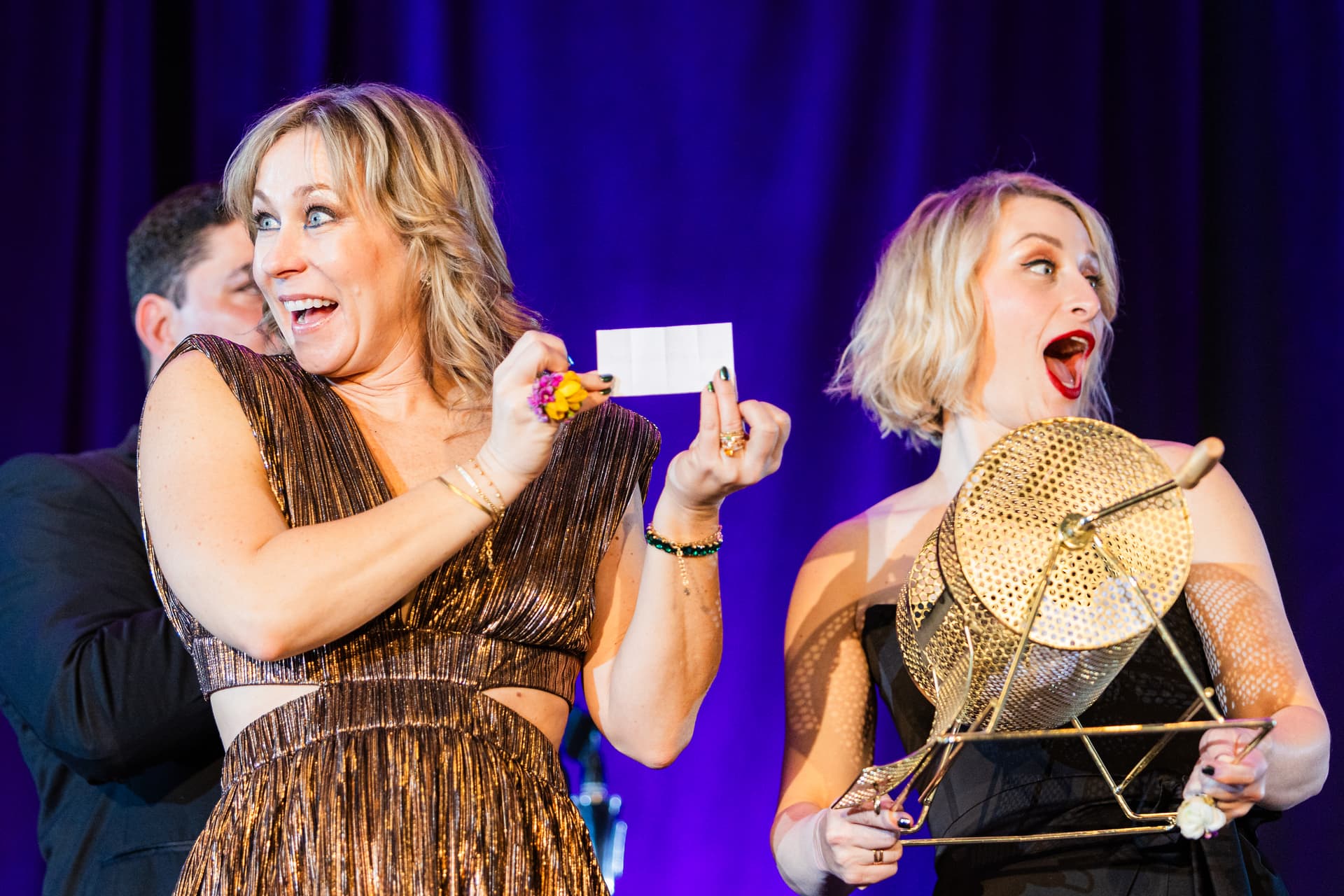 Event photo demonstrating mastery of mixed lighting — a close up shot of two women reading a winner off a piece of paper, with warm lighting and purple backlight