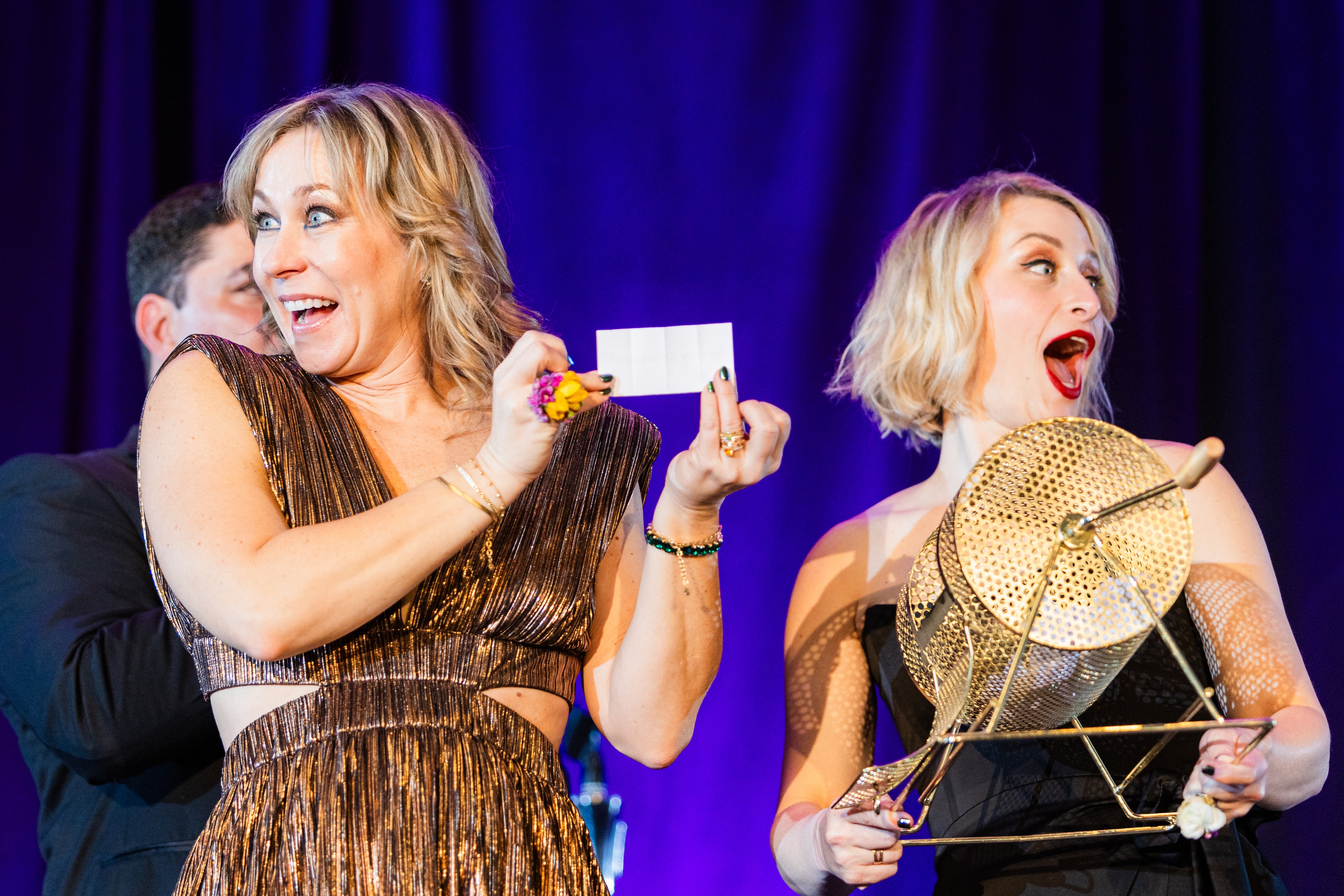 Event photo demonstrating mastery of mixed lighting — a close up shot of two women reading a winner off a piece of paper, with warm lighting and purple backlight