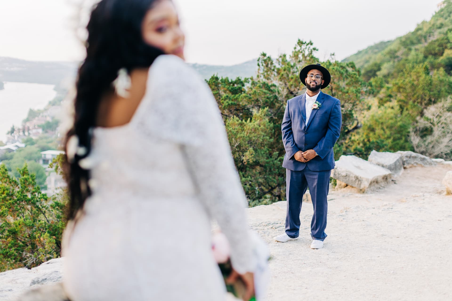 A groom looking at his bride in the foreground as she looks over her shoulder towards the camera. They stand on a clearing on Mt. Bonnel so you can see Lake Austin & greenery behind them.