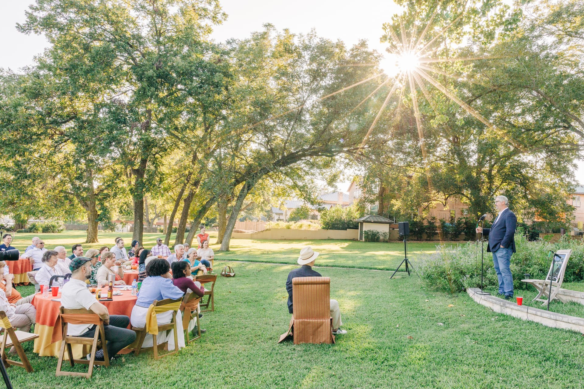 Milestone birthday toast — a large party on a lawn during golden hour as a toaster gives a speech about the birthday celebrant