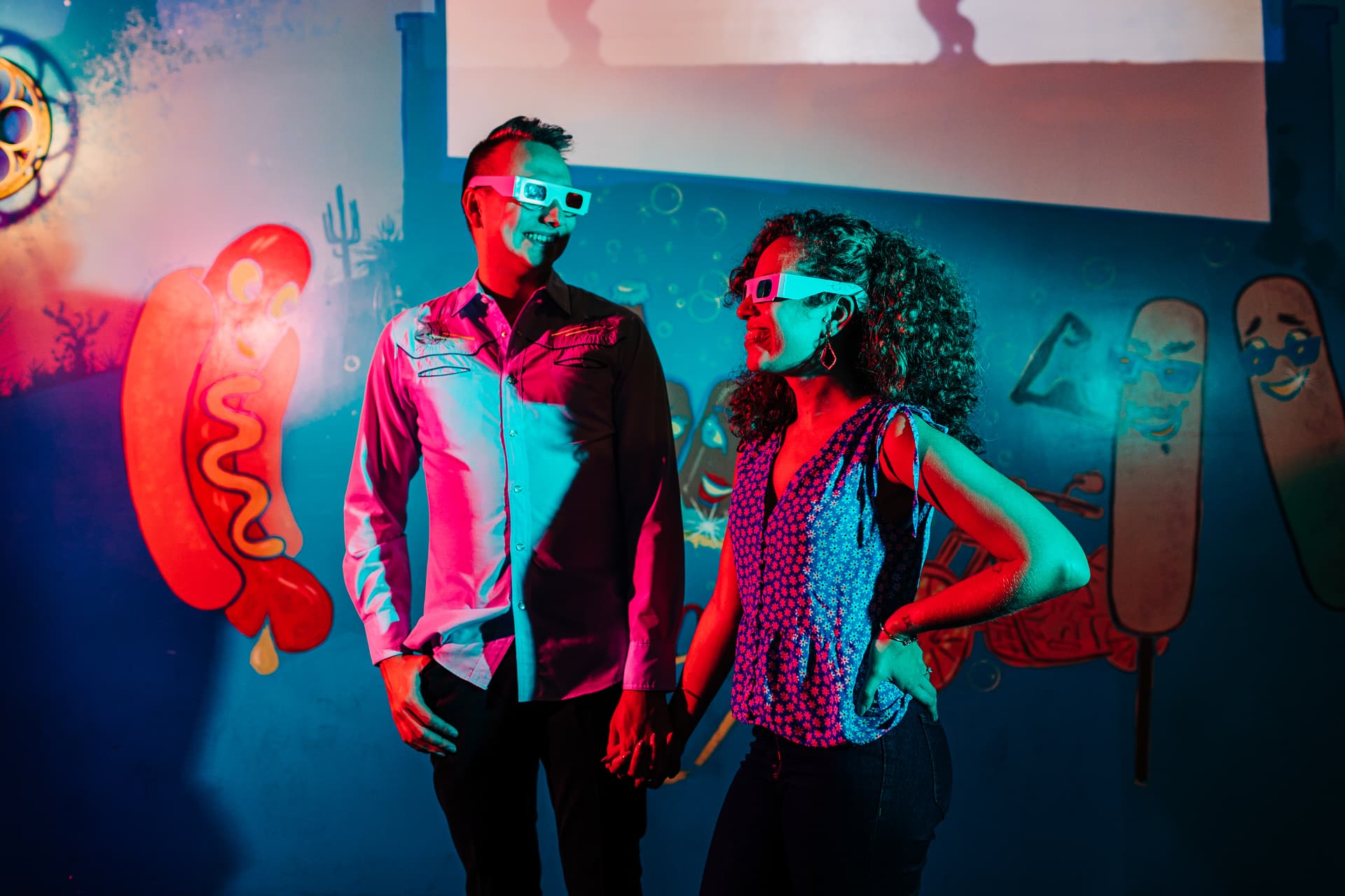 a couple in dramatic red and blue light, wearing 3d glasses inside of a movie theater with dramatic posing