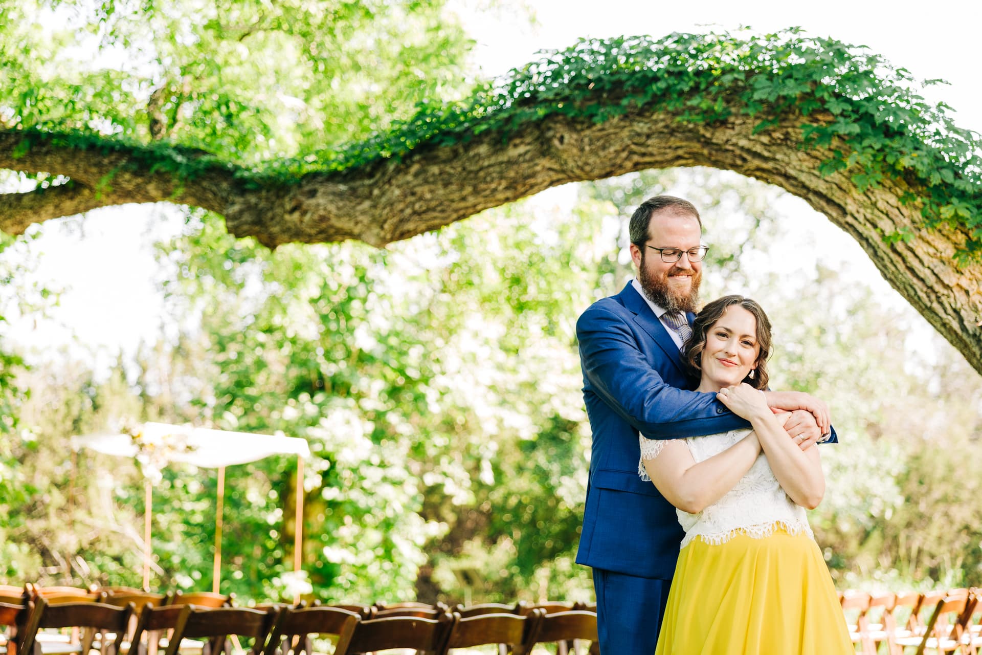 a couple with joyous expressions, posed in a warm hug in a natural environment with a large oak tree in the background. the wife is wearing an alternative yellow & white dress, and the husband is embracing her softly.