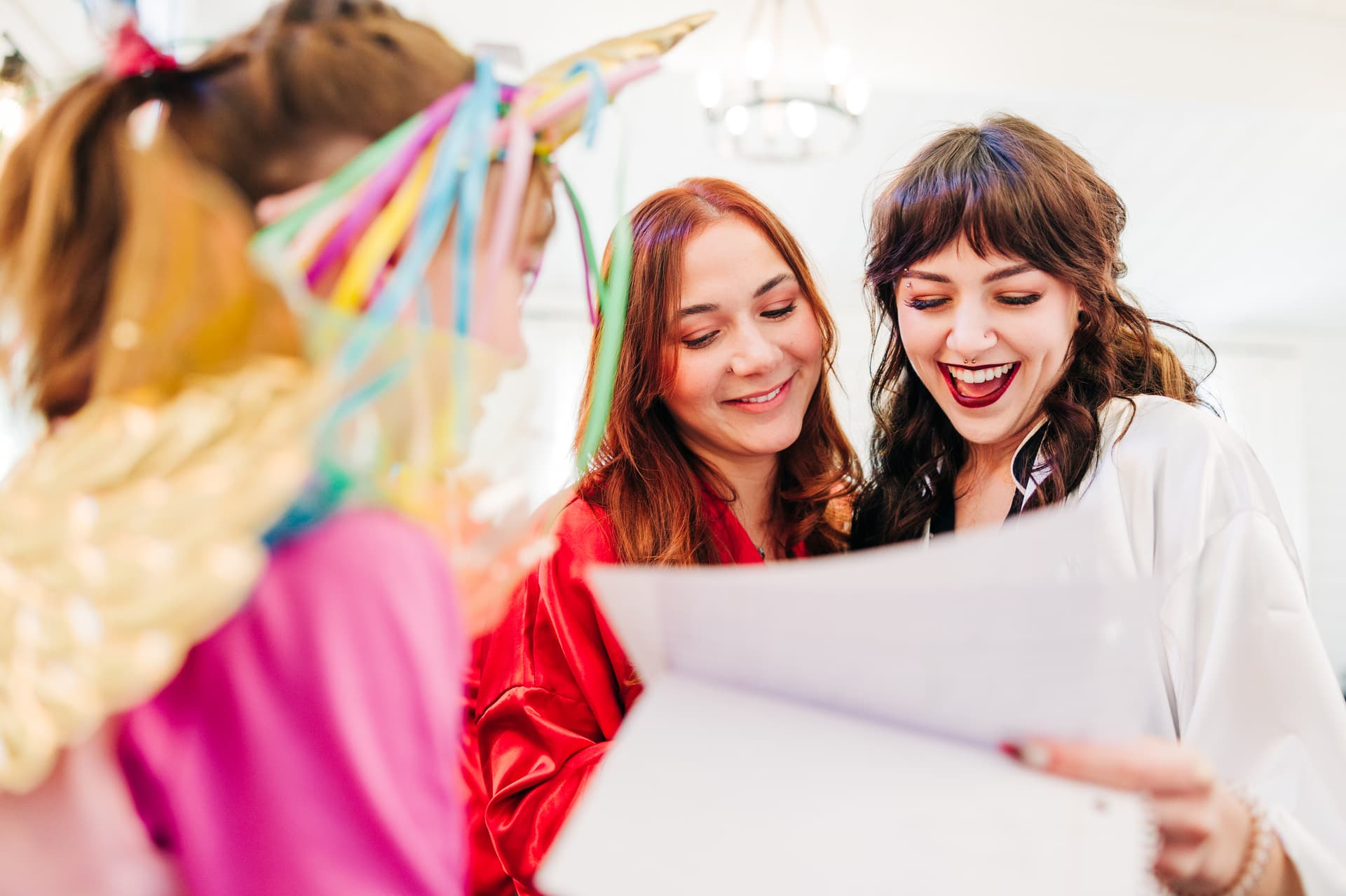 a warm moment between a bride and her maid of honor as they read a note written by her flower girl. both have joyful expressions with the flower girl in the foreground, and look excited by what they are reading.