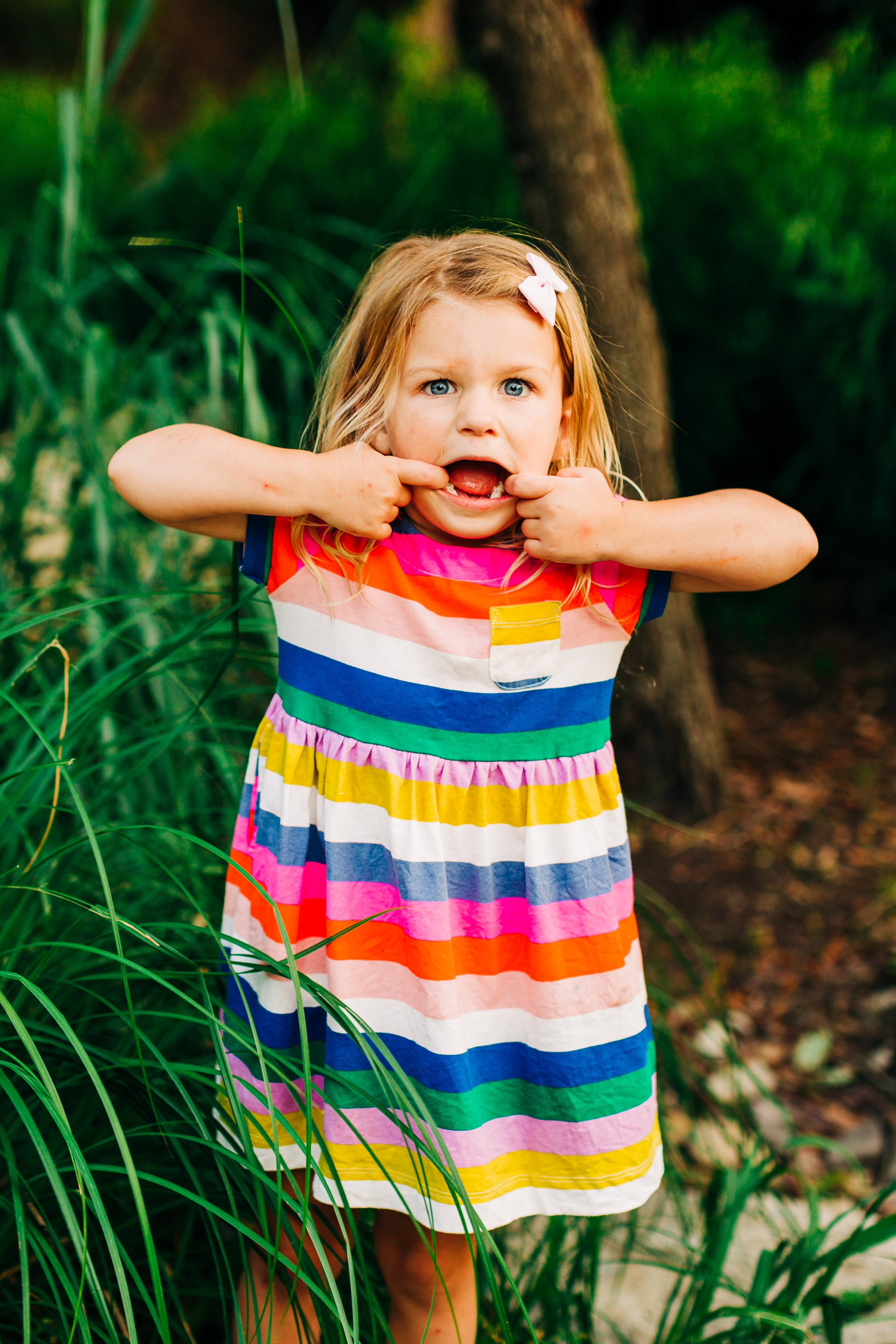 Young child makes a silly face at the camera, pulling her mouth out to the sides pretending to be a monster. She stands near tall grass wearing a bright colorful dress.